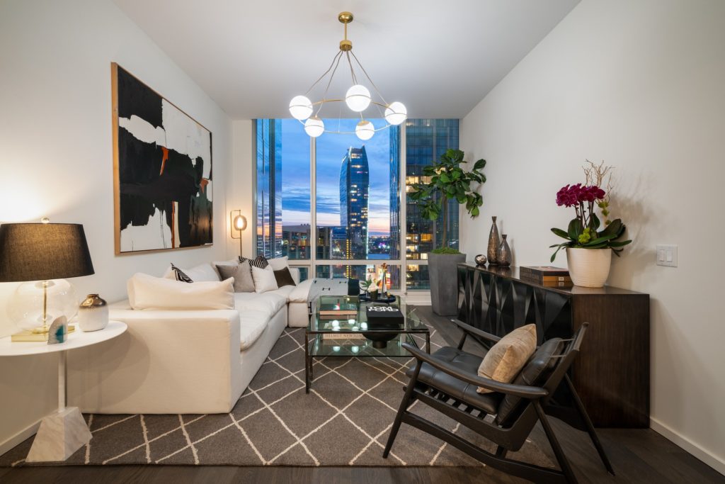 Studios in Downtown Los Angeles, CA - THEA at Metropolis - Living Room with Couch, Table, TV Stand, Rocking Chair, Chandelier, and Large Window with City Views