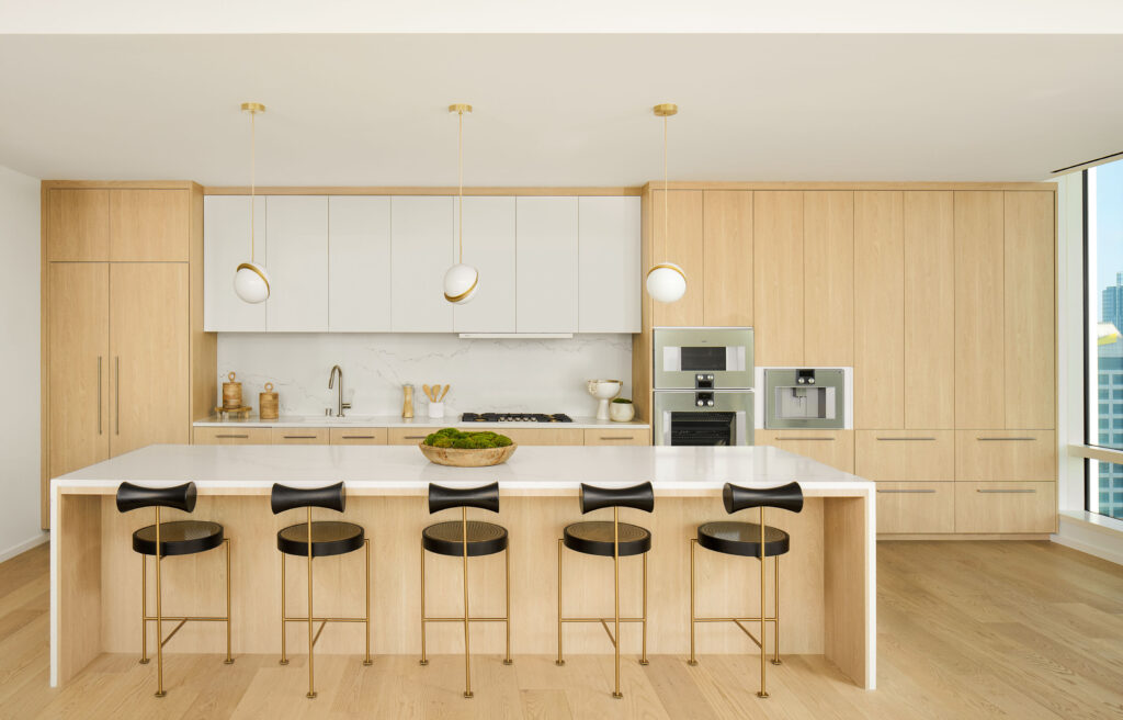 Kitchen with long white countertop with black barstools and white and wood cabinetry with gray appliances and white and gold lighting