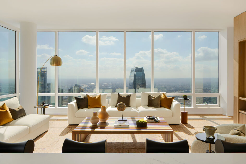 Living room with white couch and loveseat around a centered wooden coffee table with brown and green accents against floor-to-ceiling windows showing city views.